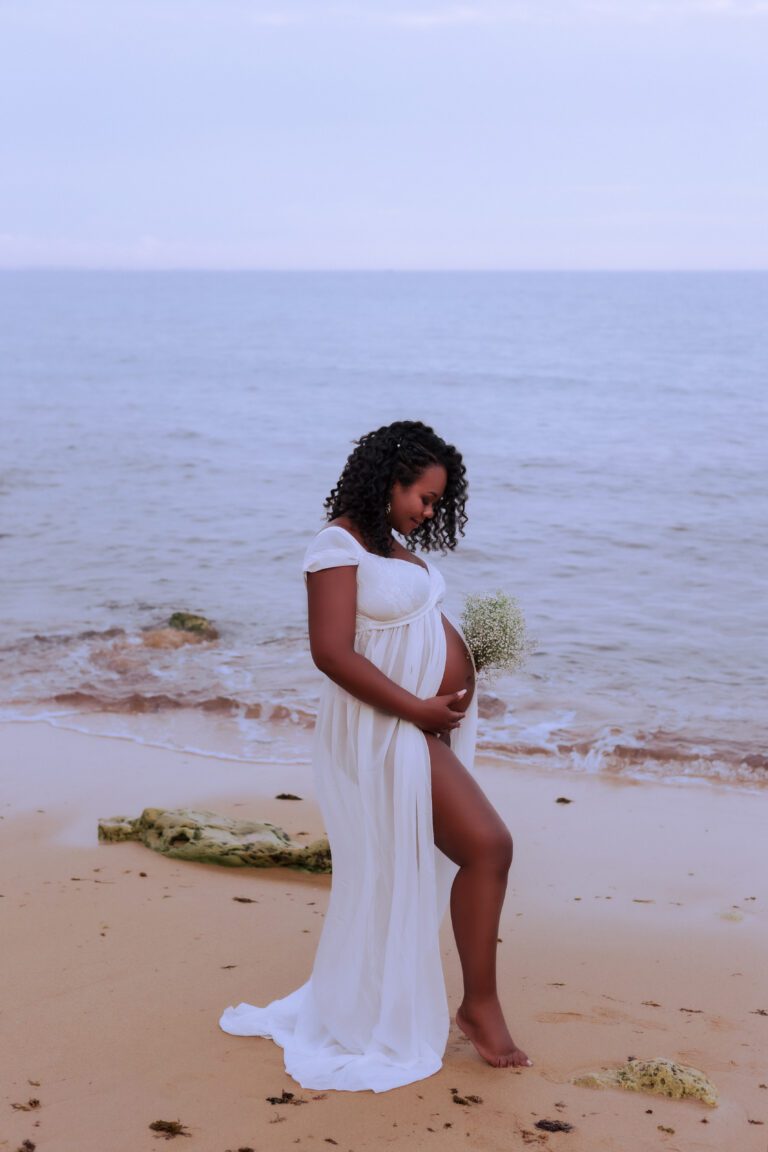Grávida de vestido branco, de perfil junto à água numa praia.
