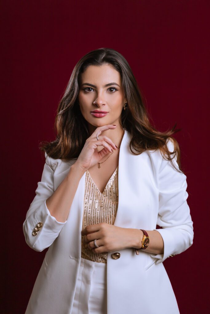 Raiza Barbozza de blazer branco, com uma mão junto ao rosto, num retrato vertical.