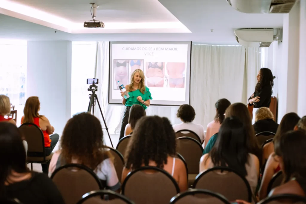 Oradora a apresentar junto a um ecrã, perante participantes sentados numa sala de evento, com câmara montada em tripé.