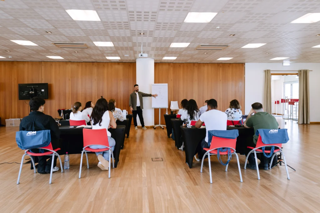 Formador junto a um flipchart, perante participantes sentados em mesas dispostas em U, numa formação de empresa.