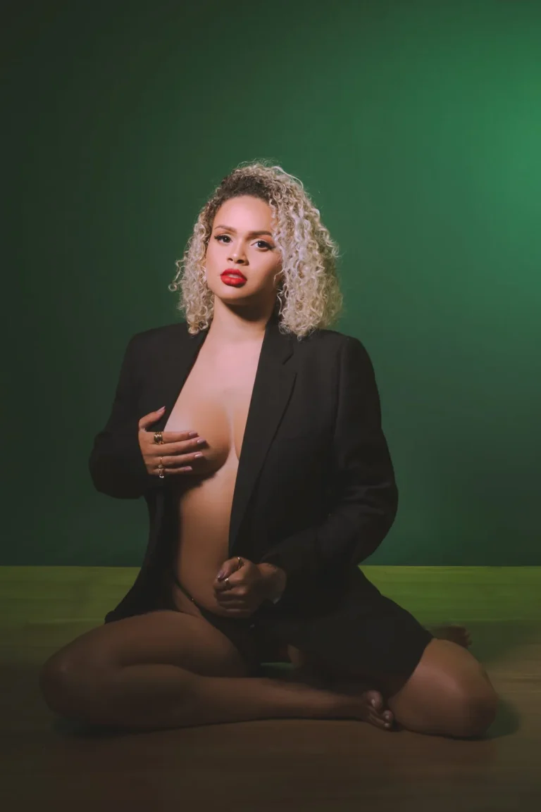 Mulher de blazer preto sentada no chão do estúdio, sobre fundo verde escuro, em sessão boudoir.