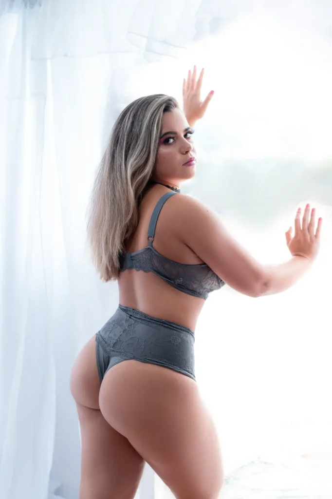 Mulher de pé junto a um cortinado branco, em lingerie cinzenta, em contraluz de janela.
