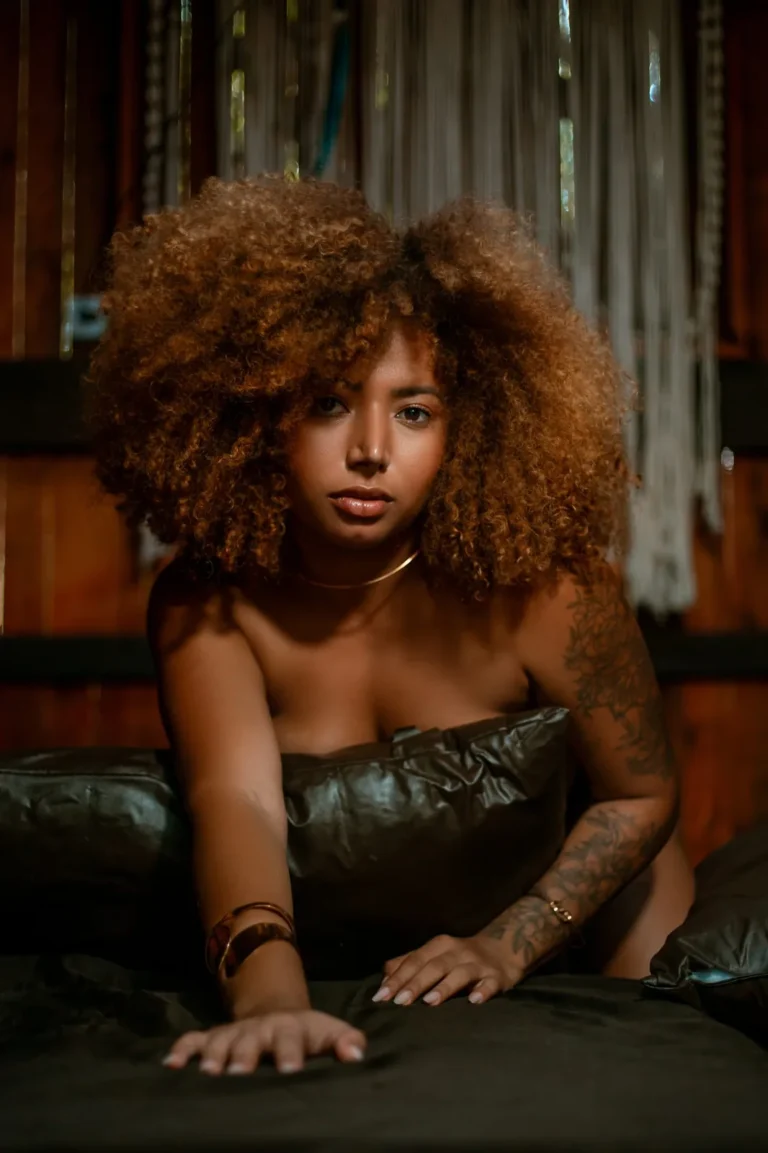 Mulher de cabelo afro apoiada num sofá escuro, com luz quente lateral, em sessão boudoir.